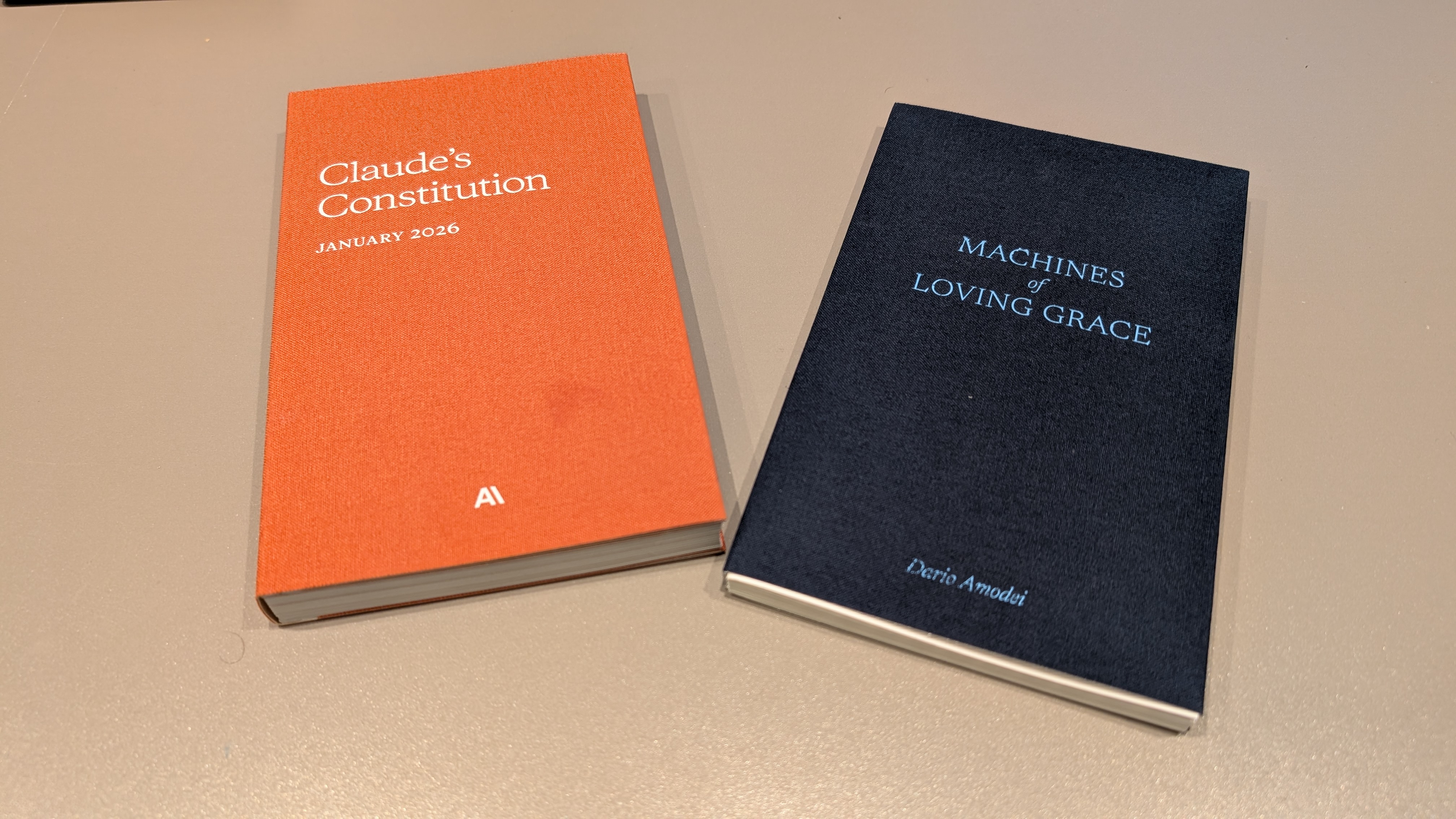 Two cloth-bound books handed out at the convening: Claude's Constitution (January 2026) in orange, and Machines of Loving Grace by Dario Amodei in navy.